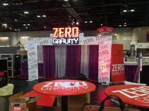 Trade show booth with a "Zero Gravity" theme features LED signage above a curtain, creating a captivating glow. The tables and walls are adorned with the brand's logo and text, while various boxes and equipment are scattered around, enhancing the immersive experience.