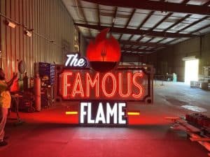 Inside a spacious warehouse, a large custom sign reading "The Famous Flame" glows brilliantly. Featuring a red flame with a fork design above the text, this striking piece of custom signage casts its vibrant glow throughout the dimly lit space.