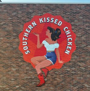 A brick wall mural depicts a woman in a white off-shoulder top, blue shorts, and red boots, sitting with one leg crossed and holding a drink or pointing upwards. The words "Southern Kissed Chicken" encircle her on a red background. Created by Sign Company Orange County, this artwork adds flair to the space.