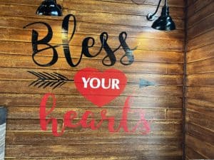 A wooden wall features a decorative sign from a renowned Sign Company in Orange County that reads "Bless Your Hearts." The words "Bless" and "hearts" are in stylish cursive font, with "Bless" in black and "hearts" in red. The word "YOUR" is in white on a red heart shape, with two arrows pointing outward from the heart.