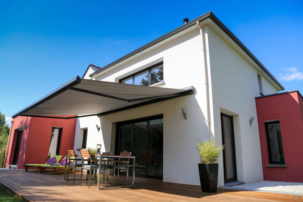 Modern two-story house with white and red exterior, large windows, and a retractable awning over a wooden deck. The deck is furnished with an outdoor table, chairs, and lounge chairs. Bright blue sky and green shrubbery add to the inviting ambiance, provided by our friends at Custom Sign Services Orange County.