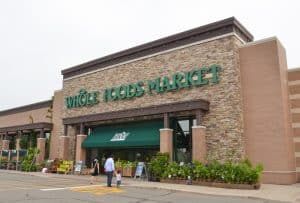 A Whole Foods Market storefront features a brick and stone facade with a green awning from Custom Sign Services Orange County. Several plants are displayed outside the entrance. Customers, including a person and a child, are walking towards the store. The sky appears overcast.