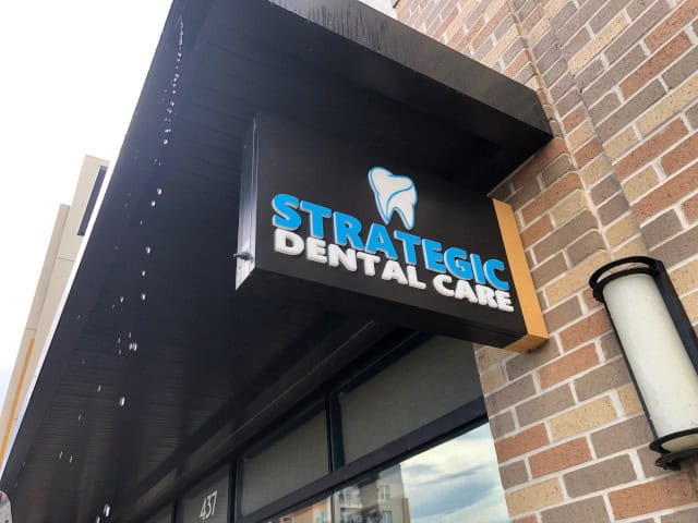 A sign for "Strategic Dental Care" hangs above a doorway, created by a top-tier Sign Company in Orange County. The sign features a tooth graphic and brightly colored text. The building exterior is made of brick, with another structure partially visible. A light fixture is mounted on the wall.