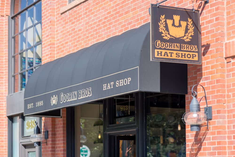 A brick building with a black awning reads "Goorin Bros Hat Shop," showcasing their custom sign services in Orange County. A matching black sign with a gold emblem hangs nearby. The shop has large windows and an antique-style exterior light fixture beside the entrance. The year "Est. 1895" is displayed on the awning.