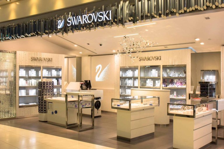A Swarovski store featuring a modern interior with illuminated display cases presenting various crystal products. The store, enhanced by a chandelier and eye-catching business signs from Orange County, proudly showcases the Swarovski logo and swan emblem, creating an elegant shopping environment.
